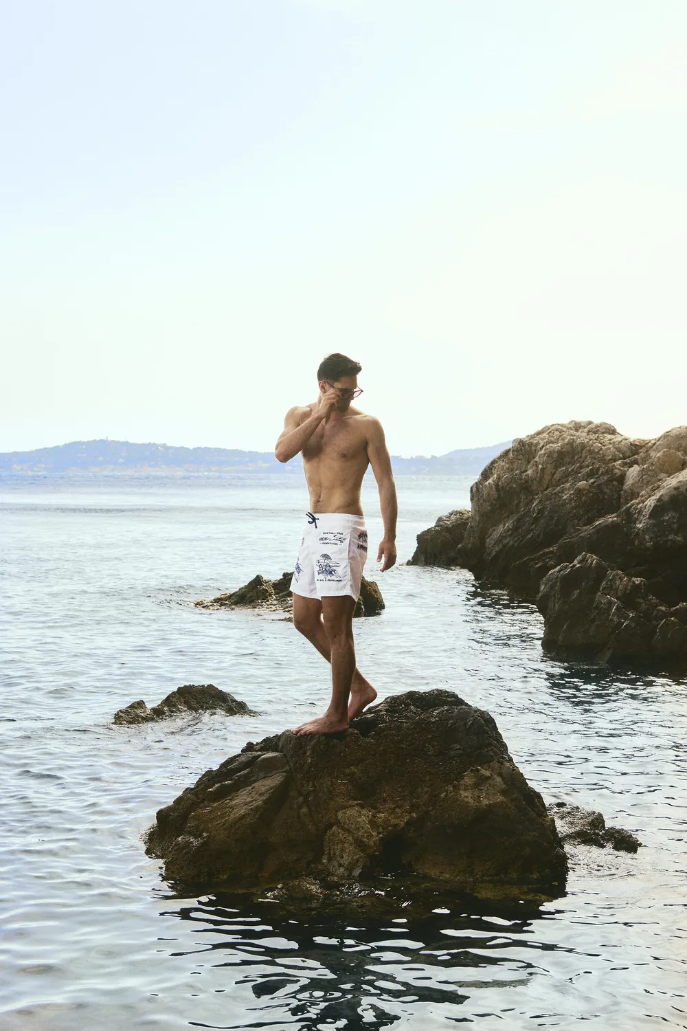 A man in white printed swim shorts stands on a rock in open water. The print on the shorts is a drawn postcard: Greetings from Bain de Mer, French Riviera.