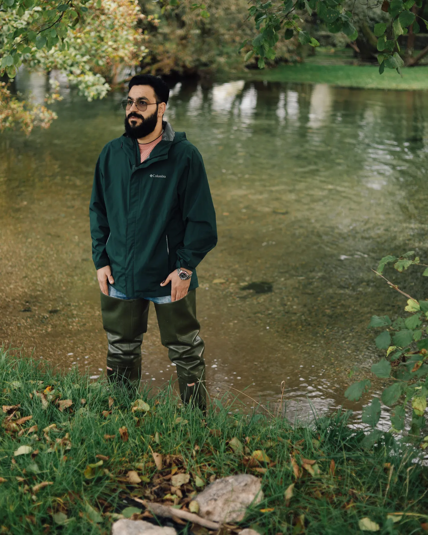 Suleiman standing in shallow river water in a green jacket and waders, looking out of frame, from the By Us, For Us campaign