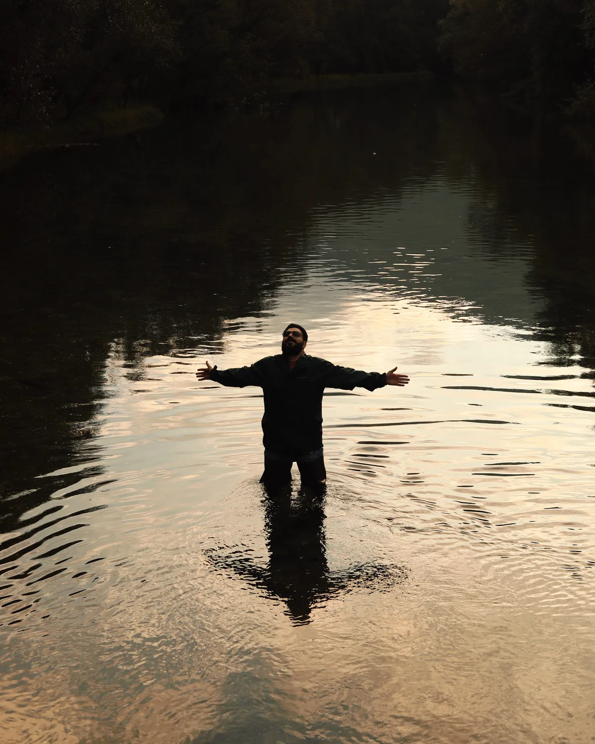 Suleiman standing in a river at dusk, arms open, from the By Us, For Us campaign