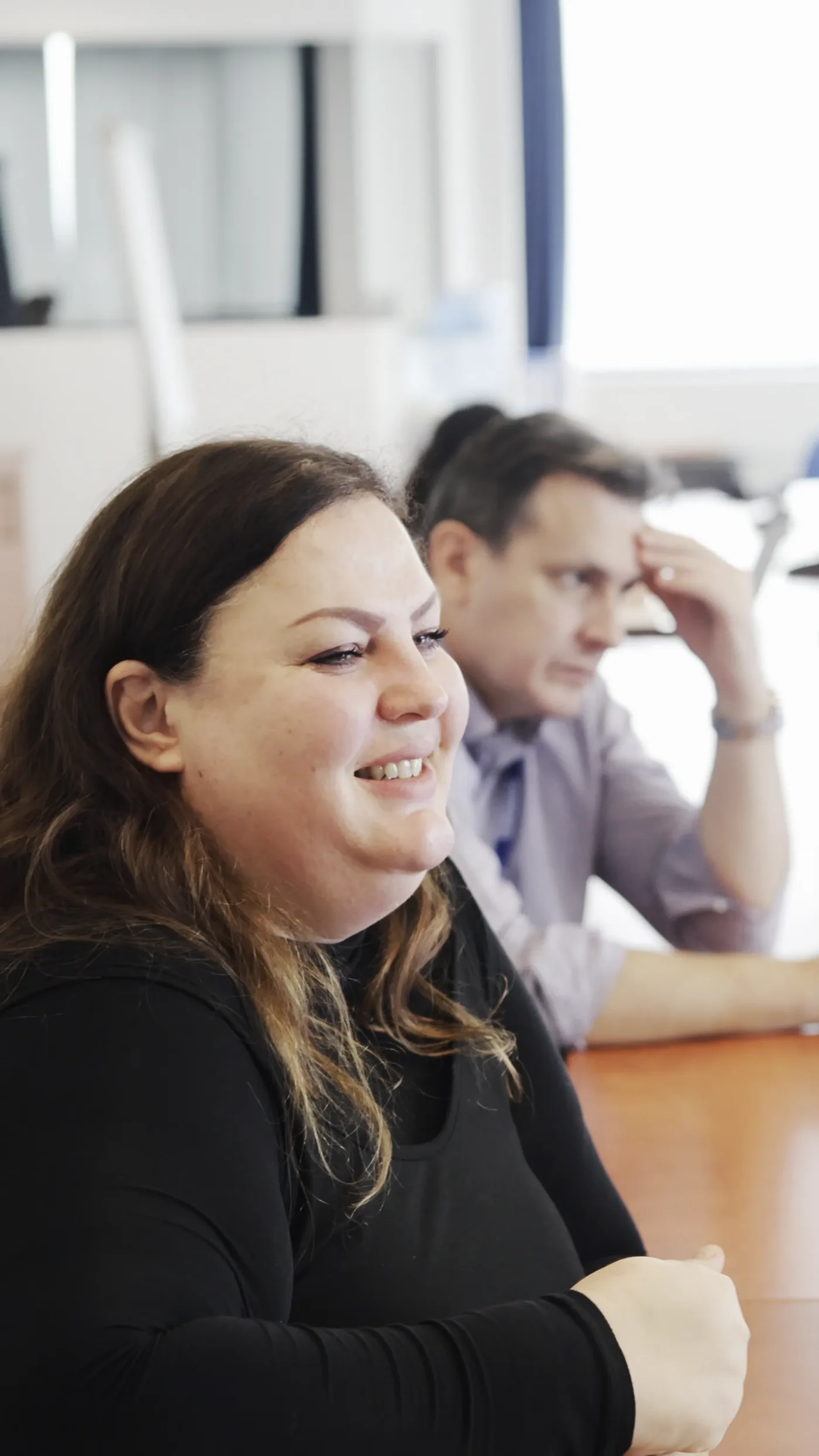 A woman smiles while listening, a man behind her rests his head on his hand.