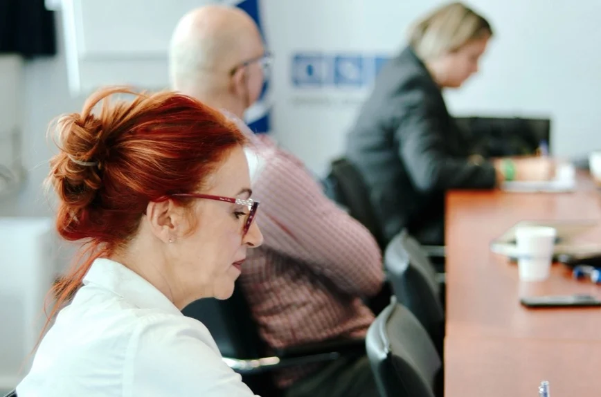 A woman with red hair writes in a notebook while others sit further along the table.