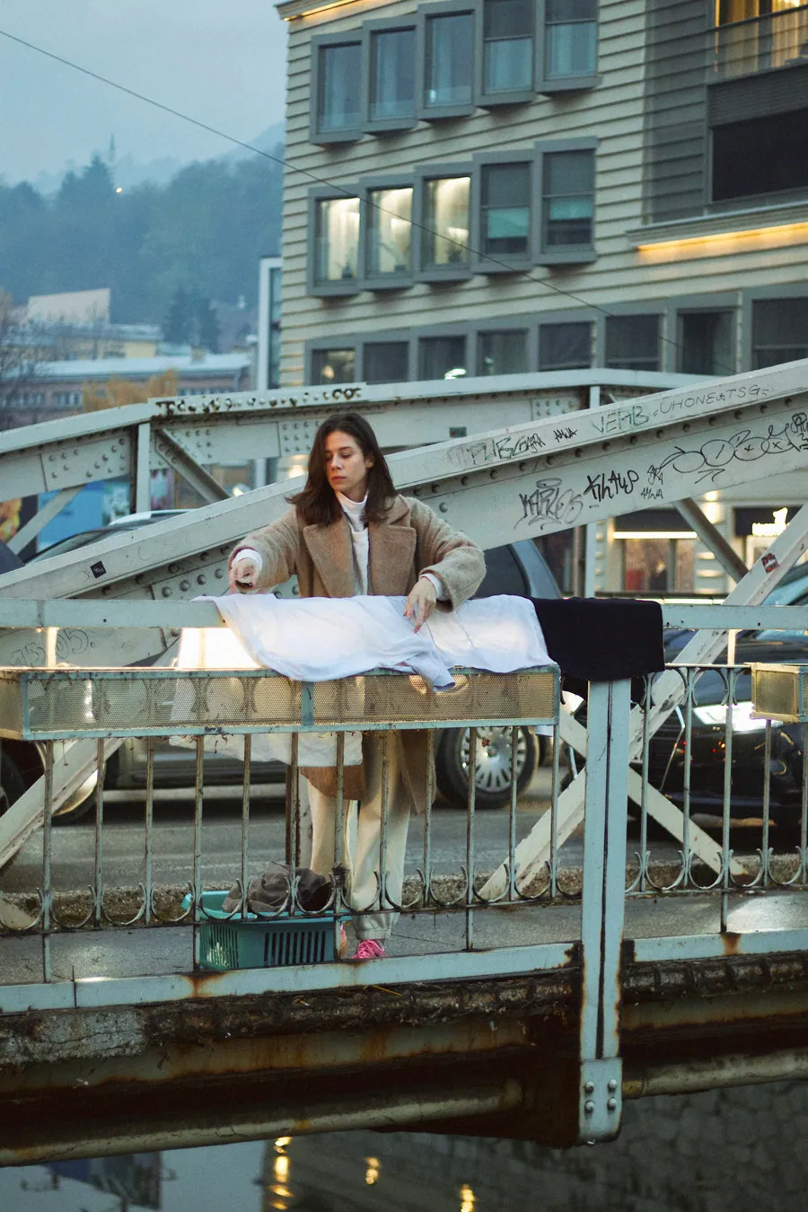 A woman stands behind a bridge railing over which white linen hangs, a teal basket at her feet, with city buildings and hills behind.