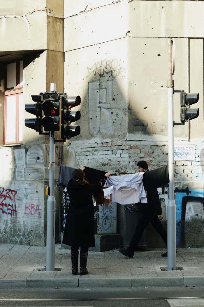A woman in black stands under a line of washing on a street corner, a traffic light beside her.