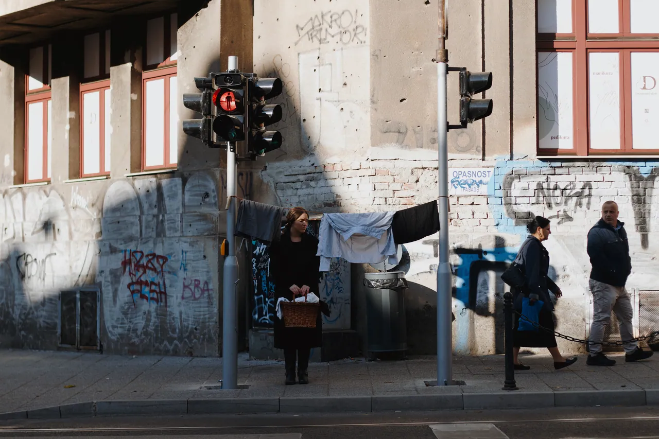 A woman in black stands beneath a line of washing strung across a street corner beside a traffic light, two men walking past on the right.