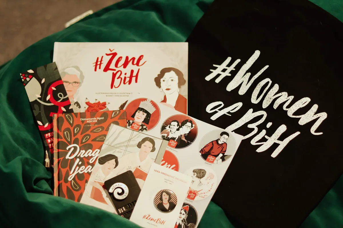 Books, cards and a black bag printed with #Women of BiH on a green cloth.