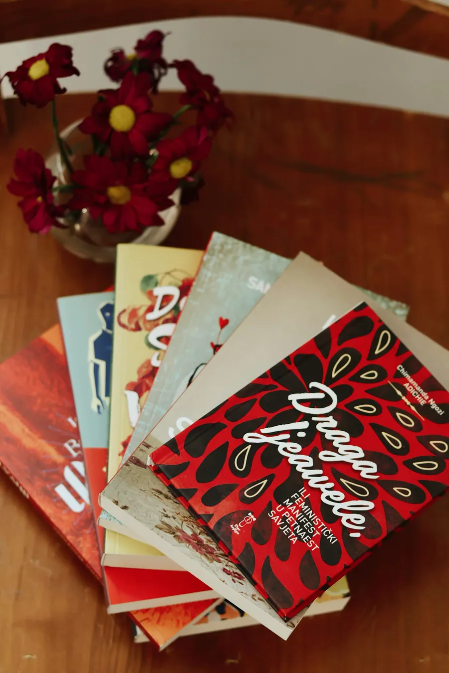 A fan of books with red covers lying on a wooden table beside red flowers.
