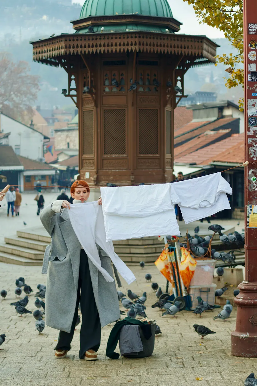 In front of the Sebilj fountain in Sarajevo, a woman holds up a white sheet beside laundry hung on a line.