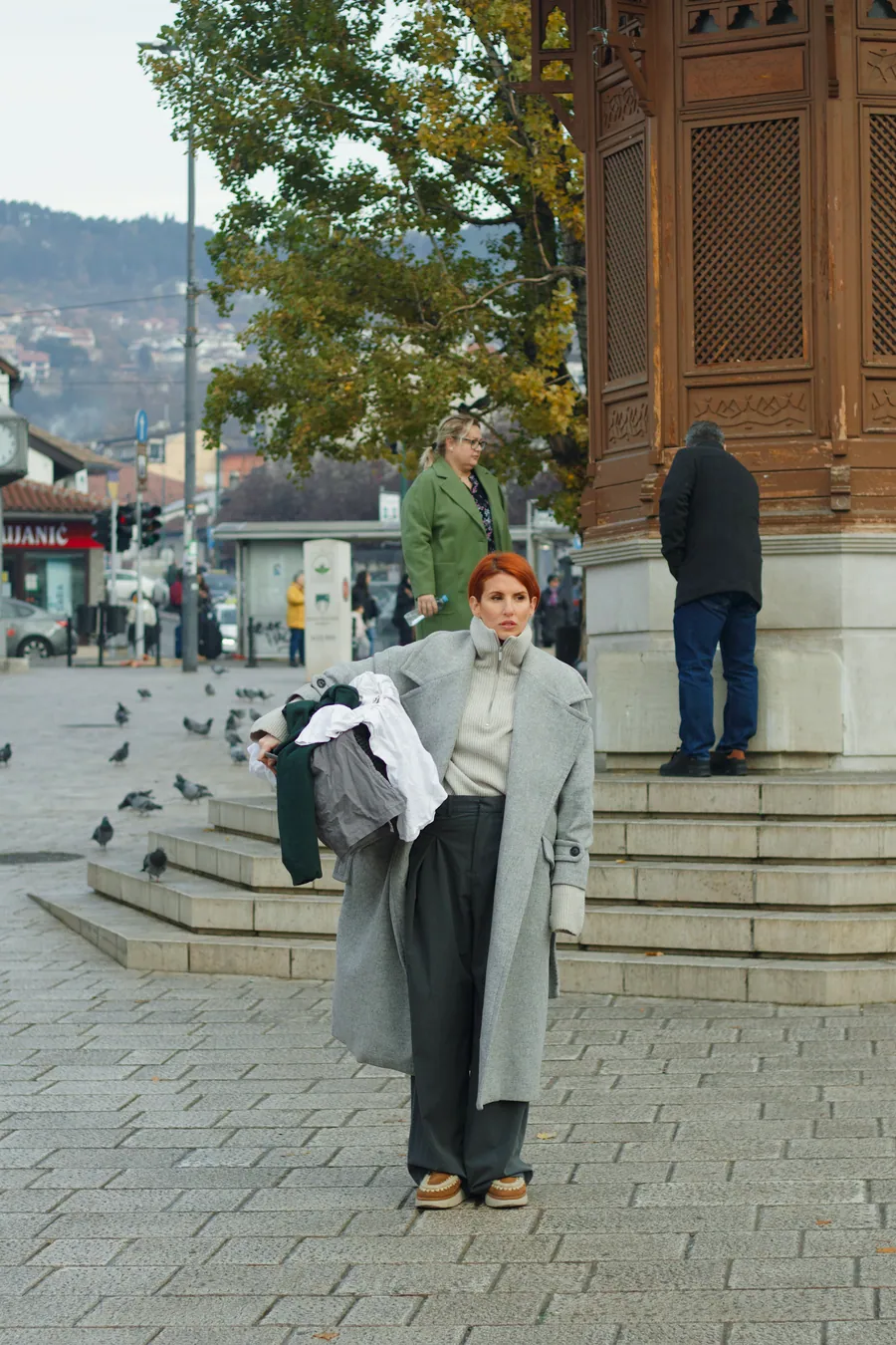 A woman in a long grey coat on the steps by the Sebilj, a bundle of dark cloth in her arms.