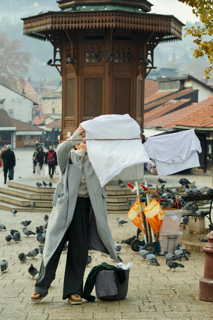 A woman in a long grey coat holds a white sheet up in front of her face at the Sebilj, pigeons around her.