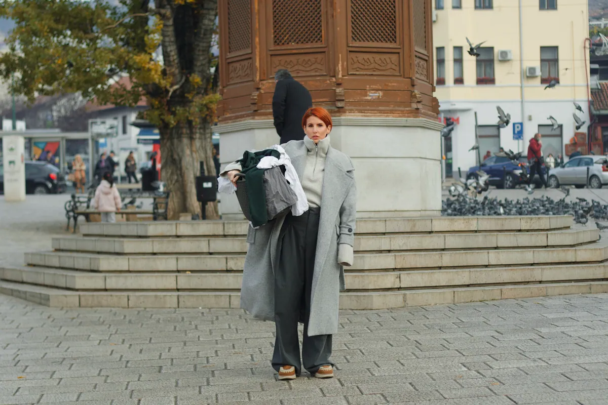 A woman in a long grey coat stands on the steps by the Sebilj fountain holding a bundle of dark cloth, a bare tree and the square behind her.