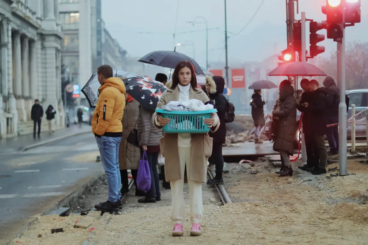 A woman stands at a street crossing holding a teal laundry basket, in front of a group of pedestrians with umbrellas, a red traffic light behind them.