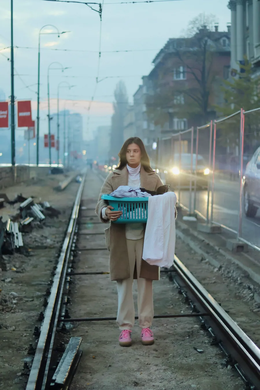 A woman stands between tram tracks in the early light, a teal basket in her arms, the street behind her.