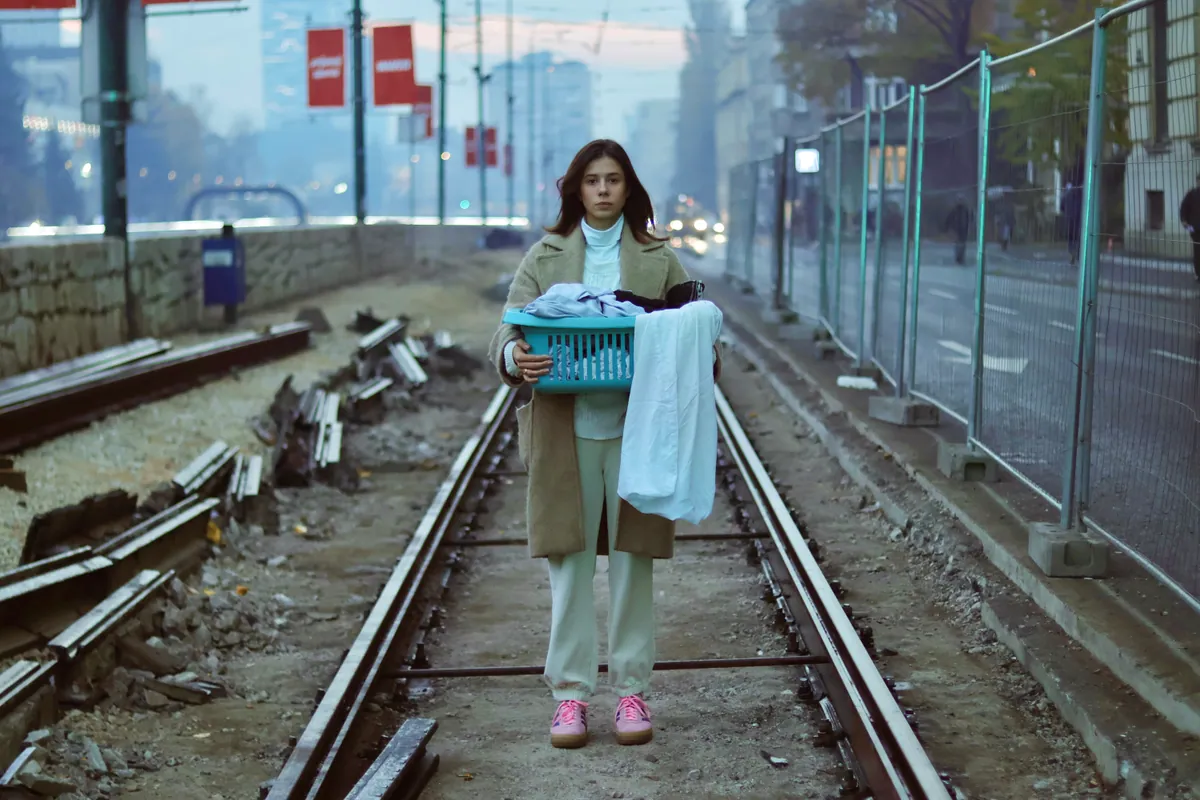 A woman in an olive coat stands between tram tracks holding a teal basket of white linen, red banners on lamp posts along the street behind her.