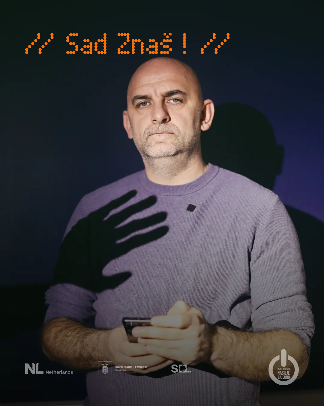 A man looks straight at the camera in a dark room, the shadow of a hand falling across his chest.
